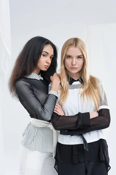 Mulheres multiétnicas atraentes em roupas pretas e brancas em pé e olhando para a câmera juntos isolados em branco — Fotografia de Stock