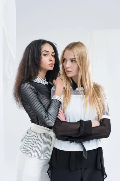 Atractivo multiétnico rubia y morena mujeres en negro y blanco ropa posando aislado en blanco - foto de stock