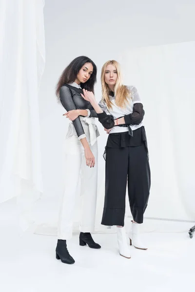 Longitud completa de atractivas mujeres multiétnicas en ropa blanca y negra posando sobre blanco - foto de stock