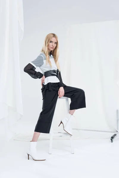 Beautiful caucasian blonde woman in fashionable black clothes putting leg on chair isolated on white — Stock Photo