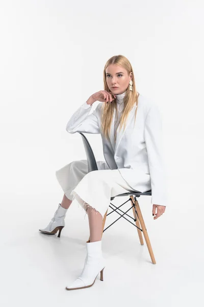 Attraente donna bionda caucasica in abiti bianchi alla moda seduta sulla sedia e guardando lontano isolato su bianco — Foto stock