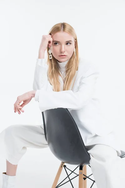 Atractiva mujer rubia caucásica en ropa blanca con estilo sentado en la silla y mirando hacia otro lado aislado en blanco - foto de stock