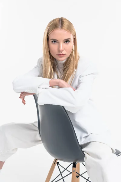 Atractiva mujer rubia caucásica en ropa blanca con estilo sentado en la silla y mirando a la cámara aislada en blanco - foto de stock