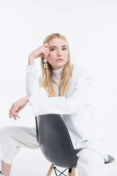 Attractive caucasian blonde woman in fashionable white clothes sitting on chair and touching head isolated on white — Stock Photo