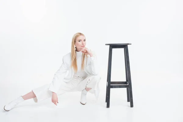 Atractiva mujer rubia caucásica en ropa blanca de moda en cuclillas cerca de silla negra aislado en blanco - foto de stock