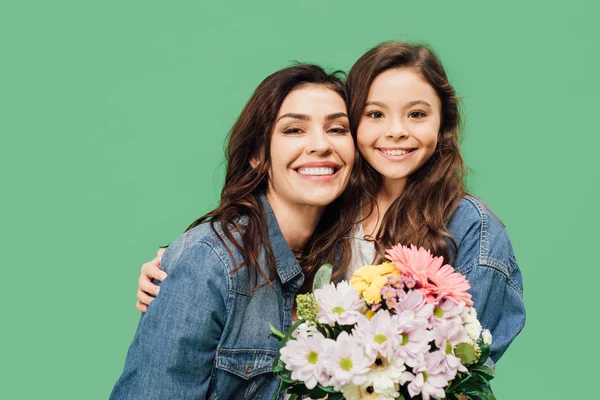 Усміхнена мати і дочка з квітами дивиться на камеру ізольовано на зеленому — стокове фото