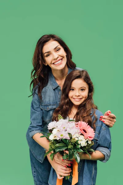 Sonriente madre abrazando hija con ramo de flores aislado en verde - foto de stock