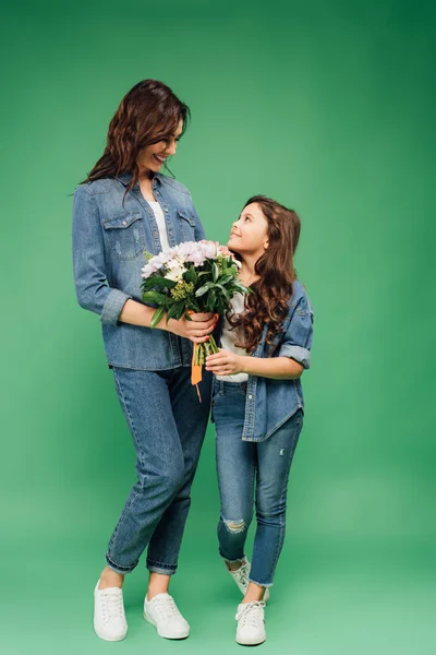 Mãe feliz e filha em jeans olhando um para o outro e segurando flores no fundo verde — Fotografia de Stock