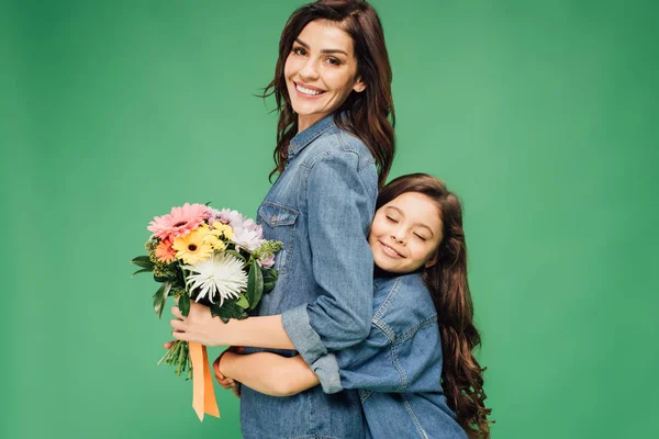 Adorable daughter hugging smiling mother with flowers isolated on green — Stock Photo