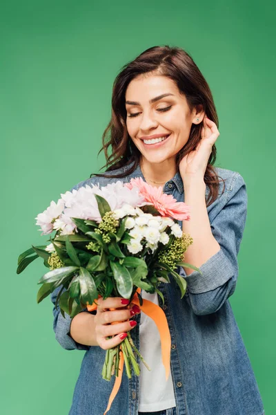 Hermosa mujer sonriente tocando el cabello y sosteniendo ramo de flores aislado en verde - foto de stock