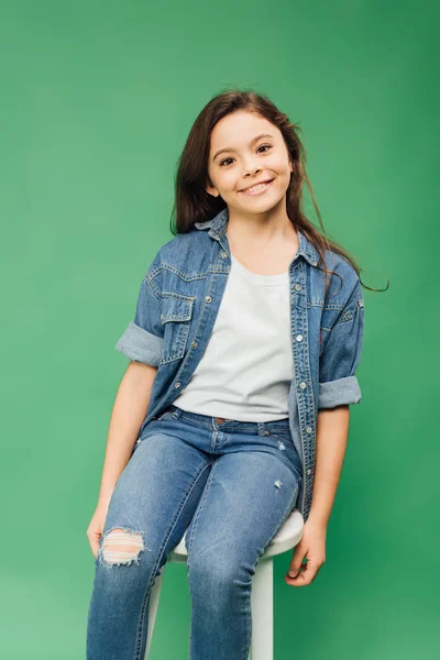 Entzückendes Kind, das auf einem Stuhl sitzt und isoliert auf grün in die Kamera schaut — Stockfoto