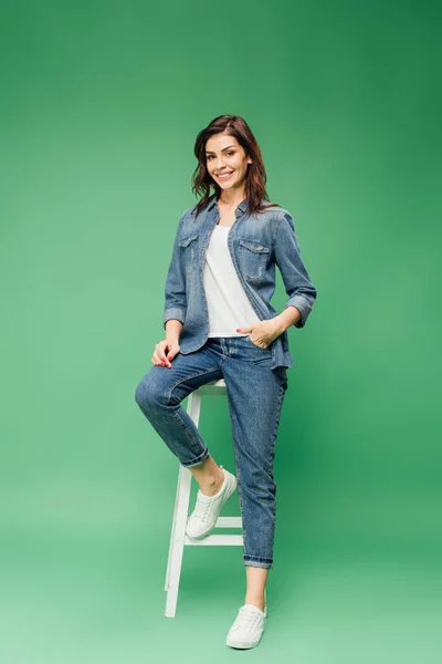 Frau in Jeans sitzt auf Stuhl und blickt in Kamera auf grünem Hintergrund — Stockfoto