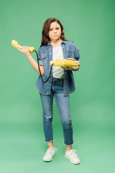 Confused child in denim holding vintage telephone and looking at camera on green background — Stock Photo