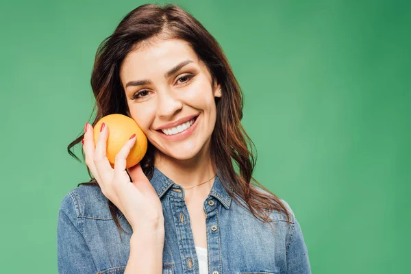 Femme souriante en denim tenant orange et regardant la caméra isolée sur vert — Photo de stock