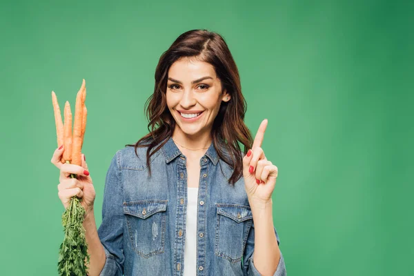 Счастливая женщина в джинсе, держащая морковь и указывая пальцем, изолированным на зеленый — стоковое фото