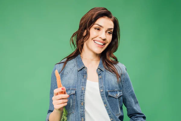 Smiling woman in denim holding carrot isolated on green — Stock Photo