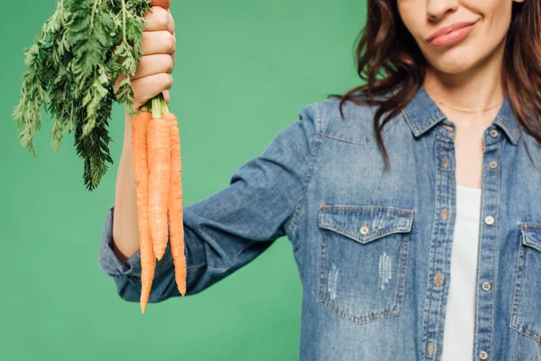 Vista recortada de mujer escéptica en denim sosteniendo zanahorias aisladas en verde - foto de stock