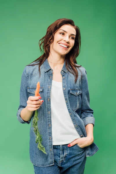 Mujer feliz en denim sosteniendo zanahoria aislada en verde - foto de stock