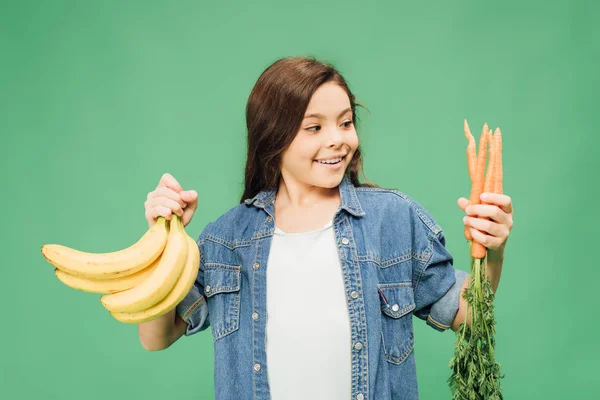 Kind hält Bananen mit Karotten isoliert auf Grün — Stockfoto