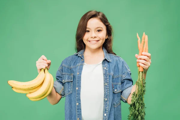 Bambino guardando la fotocamera e tenendo in mano banane con carote isolate sul verde — Foto stock