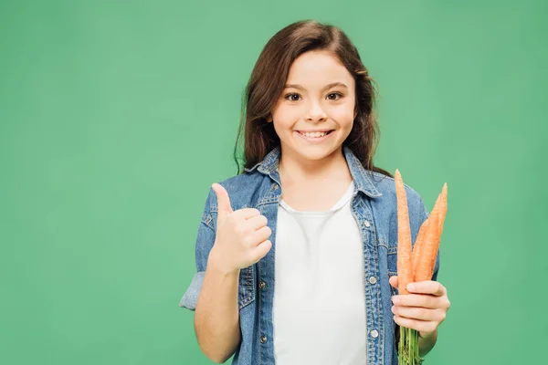 Happy child holding carrots and showing thumb up sign isolated on green — Stock Photo