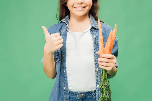 Vue recadrée de l'enfant tenant des carottes et montrant pouce vers le haut signe isolé sur vert — Photo de stock