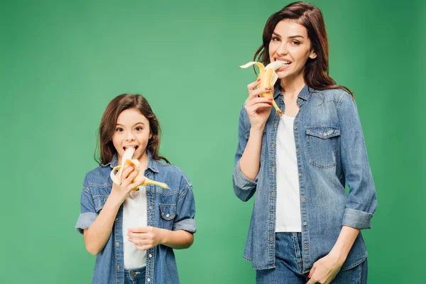 Mutter und Tochter blicken in die Kamera und essen Bananen isoliert auf grünem Grund — Stockfoto