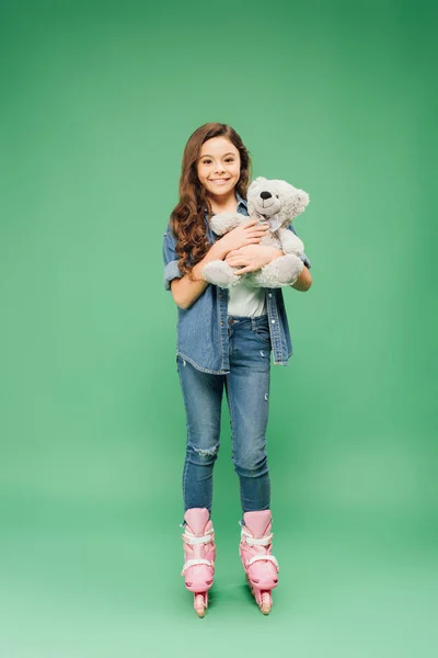 Kind in Rollerblades hält Teddybär in der Hand und blickt in Kamera auf grünem Hintergrund — Stockfoto