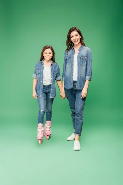 Tochter in Rollerblades hält Hand in Hand mit Mutter und blickt in Kamera auf grünem Hintergrund — Stockfoto