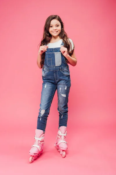 Smiling child in overalls and rollerblades looking at camera on pink background — Stock Photo