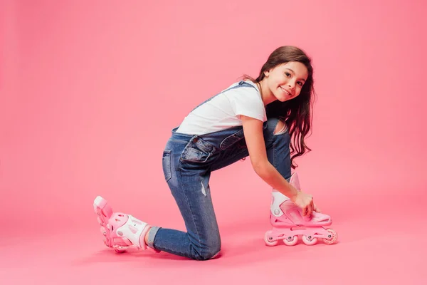 Cute child in overalls putting on rollerblades on pink background — Stock Photo