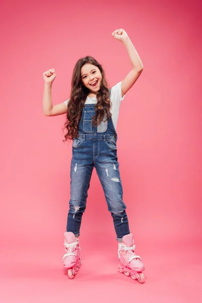 Happy child in overalls and rollerblades cheering with arms in air on pink background — Stock Photo