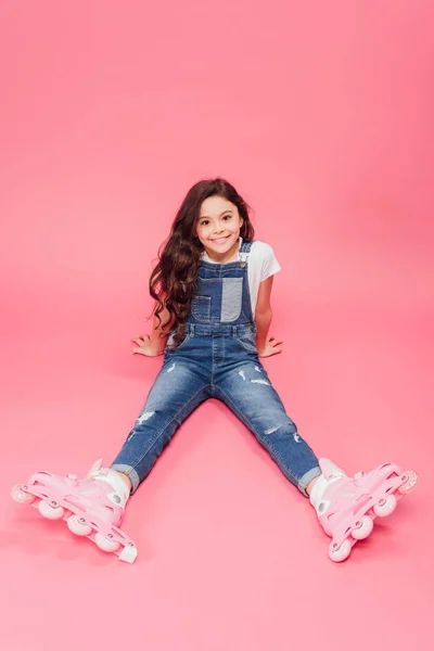 Lächelndes Kind in Overalls und Rollerblades, das auf rosa Hintergrund in die Kamera blickt — Stockfoto