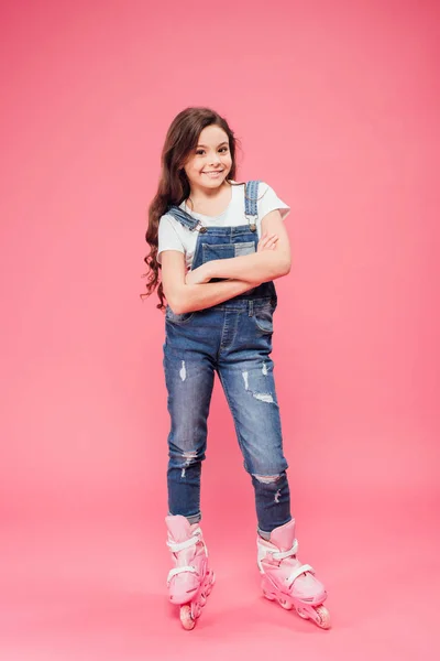 Happy child in overalls with arms crossed wearing rollerblades on pink background — Stock Photo