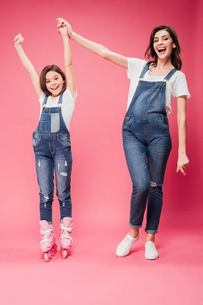 Happy daughter in rollerblades cheering and holding hands with mother on pink background — Stock Photo
