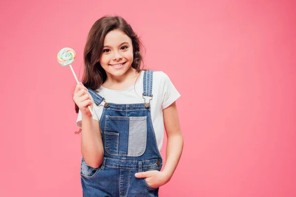 Niño alegre sosteniendo piruleta con la mano en el bolsillo aislado en rosa - foto de stock