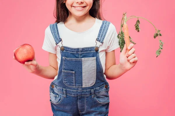 Vue recadrée du gamin tenant pomme rouge et carotte dans les mains isolées sur rose — Photo de stock