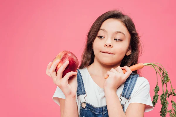 Criança olhando para maçã vermelha e segurando cenoura isolada em rosa — Fotografia de Stock