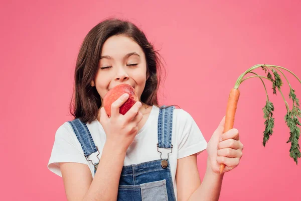 Happy child eating child red apple and holding carrot isolated on pink — Stock Photo