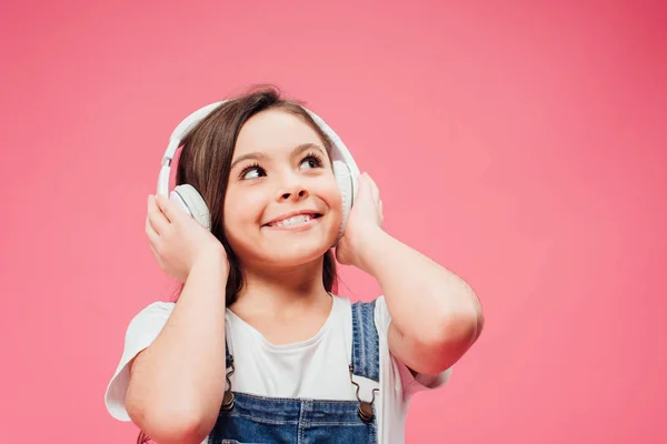 Niño alegre escuchando música y tocando auriculares aislados en rosa - foto de stock