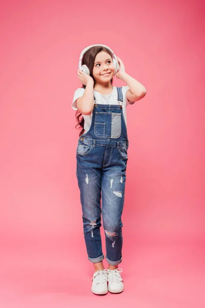 Longitud completa de niño alegre escuchando música y tocando auriculares sobre fondo rosa - foto de stock