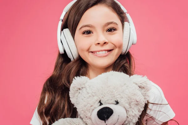 Усміхнена дитина в навушниках тримає плюшевого ведмедя ізольовано на рожевому — стокове фото