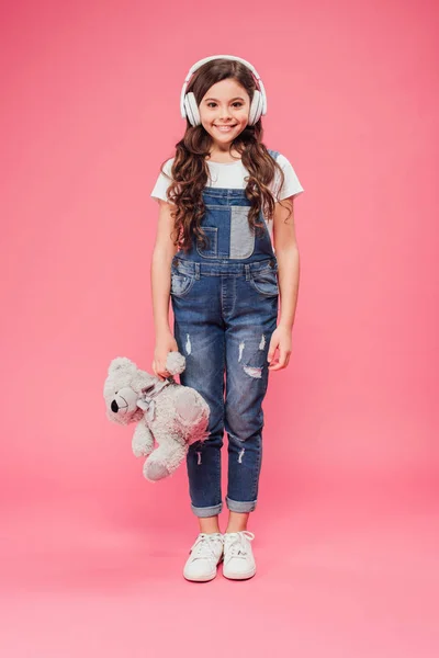Полная длина улыбающегося ребенка, стоящего в наушниках и держащего плюшевого мишку на розовом фоне — стоковое фото