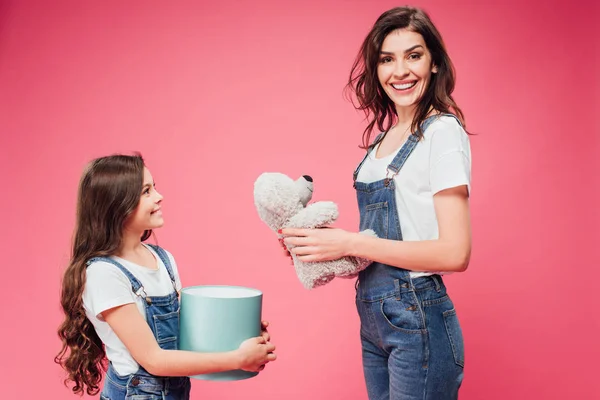 Mãe alegre com ursinho perto da filha segurando caixa de presente isolado em rosa — Fotografia de Stock