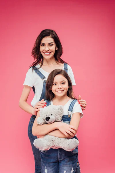 Smiling mom standing with daughter hugging teddy bear isolated on pink — Stock Photo