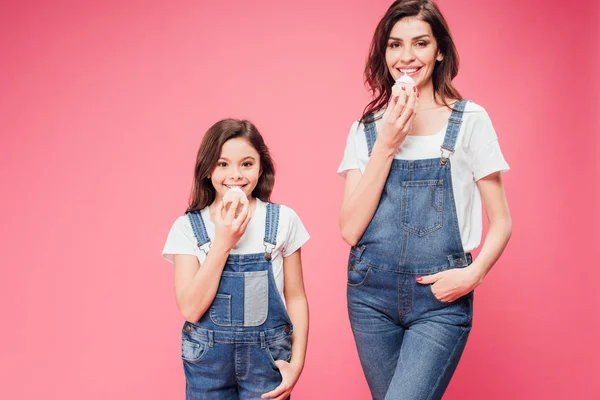 Mamá y su hija comiendo magdalenas con las manos en bolsillos aislados en rosa - foto de stock
