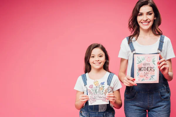 Madre e hija sosteniendo tarjetas de felicitación en el día de las madres aisladas en rosa - foto de stock