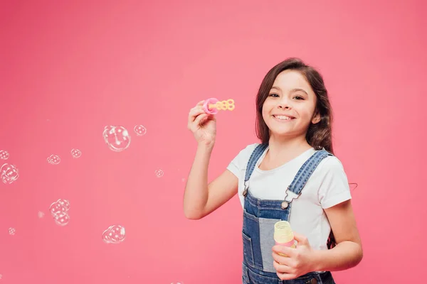 Радостный ребенок стоит рядом мыльные пузыри изолированы на розовый — стоковое фото