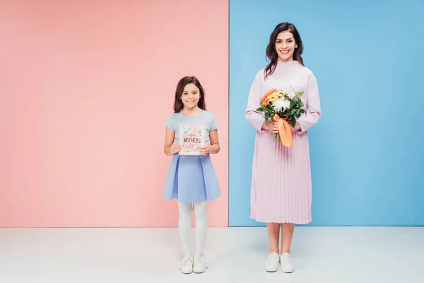 Entzückendes Kind und attraktive Frau mit Geschenken, während sie in die Kamera auf blauem und rosa Hintergrund schaut — Stockfoto