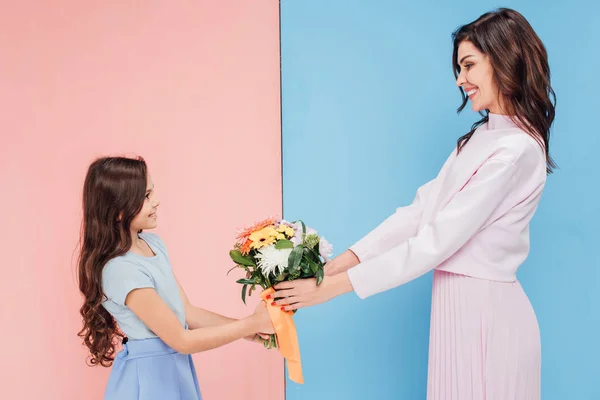 Adorável criança dando buquê de flores para mulher atraente no fundo azul e rosa — Fotografia de Stock